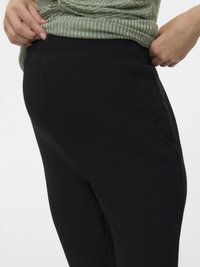 Tuvo plāna attēls, kurā persona pielāgo melno pieguļošo biksu vidukļa daļu, ģērbusies zaļā ribota krekla.