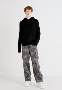 Sweatshirt preto com mangas longas e um corte descontraído, combinado com calças largas cinza em padrão camuflado e ténis brancos de sola grossa, num fundo neutro.