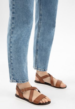 Füße in hellblauen Jeans und braunen Ledersandalen mit Knöchelriemen und weißen Sohlen auf weißem Hintergrund.