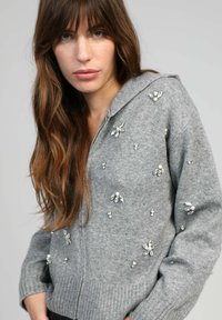 Sweat à capuche zippé gris en matériau doux, avec des embellissements décoratifs en argent et perles sous forme de motifs floraux, des poignets et un ourlet côtelés.