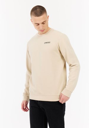 Man draagt een beige sweatshirt met het logo "PROTEST" op de borst, kijkt naar beneden, gecombineerd met een zwarte broek, staand tegen een witte achtergrond.