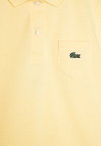 Pikétröja i mjukt, texturerat gult tyg med en treknäppt sprund, prydd med en grön broderad krokodil-logotyp på bröstfickan.