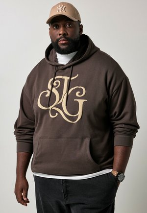 Hombre con barba que lleva una sudadera marrón con capucha y grandes letras estilizadas, una gorra de béisbol beige, pantalones negros y un reloj de pulsera, de pie frente a un fondo liso.