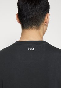 Bakifrån på en person med kort mörkt hår som bär en svart skjorta med "BOSS" i vita bokstäver vid den övre nacklinjen.