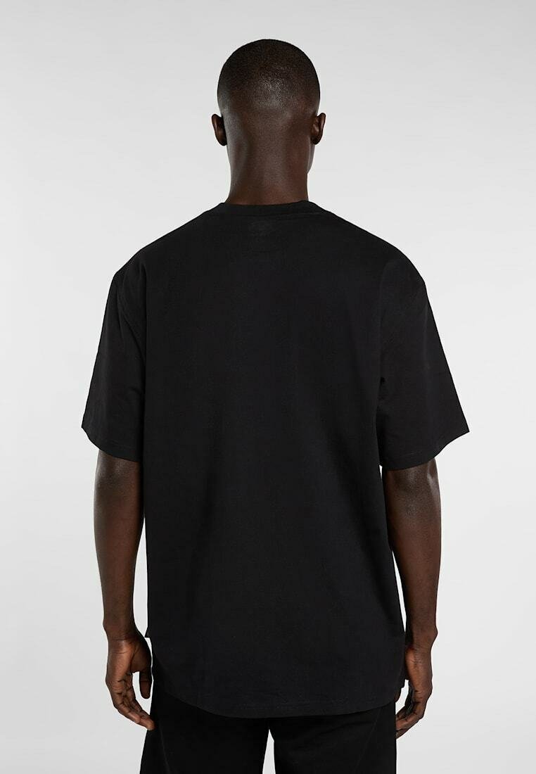 Mann mit dunkler Haut, der ein schlichtes schwarzes T-Shirt mit kurzen Ärmeln trägt, von hinten vor einem einfarbigen hellen Hintergrund gezeigt.