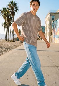 Joven caminando por un paseo marítimo iluminado por el sol cerca de una playa bordeada de palmeras, vistiendo una camiseta de rayas, jeans azul claro y zapatillas blancas.