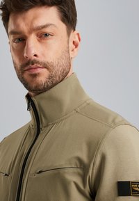 Giacca zip-up khaki con colletto alto, dotata di due tasche sul petto e un'etichetta con marchio sul maniche. Tessuto liscio, materiale leggero.