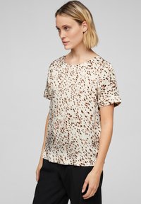 Blusa a maniche corte color crema con un motivo a macchie marroni. Presenta un collo tondo e un tessuto morbido e textured. Indossata con pantaloni neri.