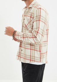 Camisa de franela a cuadros en crema, con rayas rojas y marrones, cuello abotonado y bolsillo en el pecho. Mangas largas, ajuste holgado.