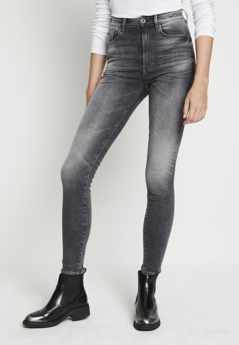Personne portant un jean skinny noir délavé taille haute et des bottines en cuir noir, debout devant un fond clair uni.