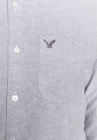 Šedá košile na knoflíky vyrobená z texturované látky, s bílými knoflíky a náprsní kapsou s vyšitou detailní podobou orla.