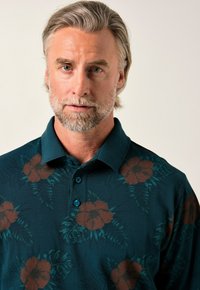 En medelålders man med grått hår och skägg, iklädd en mörk teal pikétröja med bruna blommönster mot en enfärgad bakgrund.