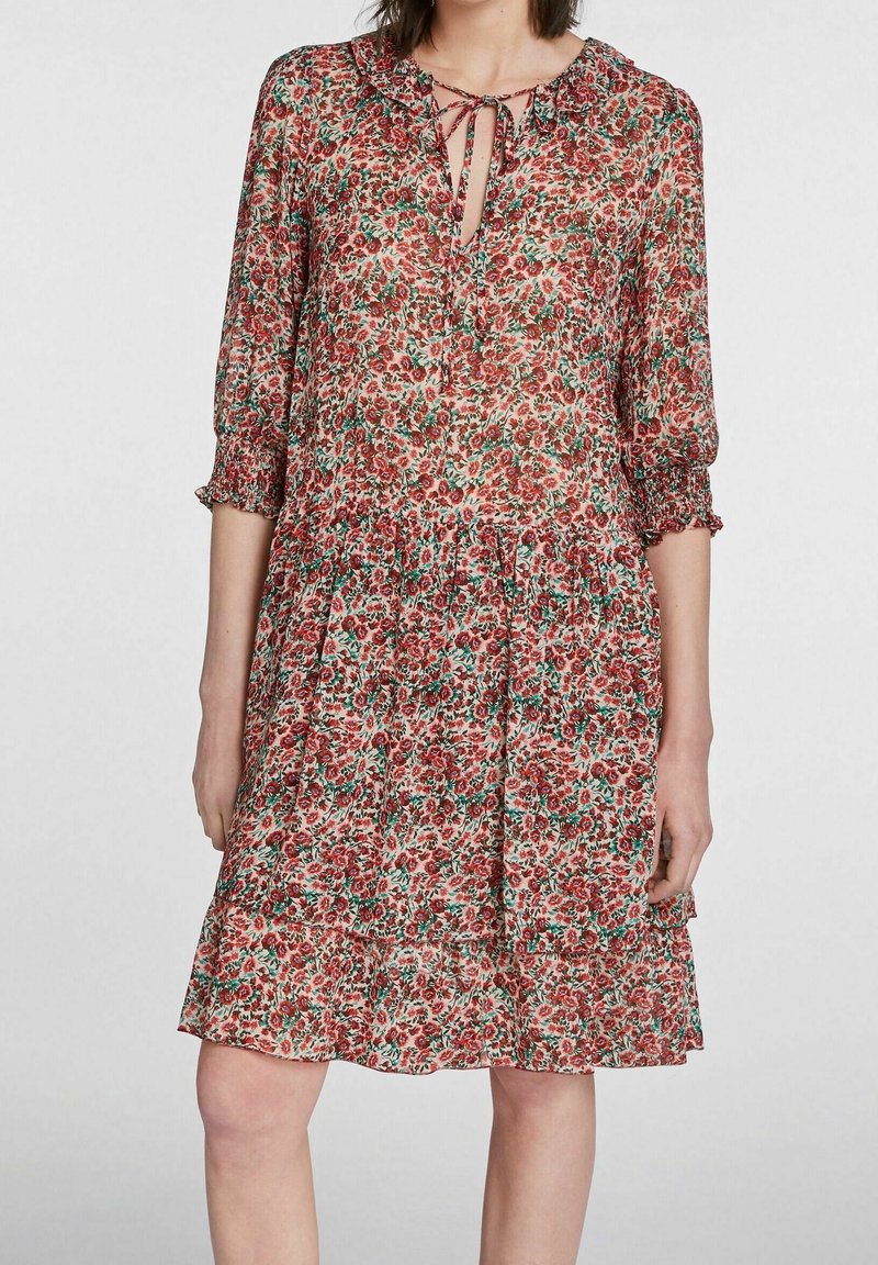 Robe fleurie avec un petit col, un décolleté en forme de trou de serrure, et des manches trois-quarts avec des poignets à volants. Motif de fleurs rouges et vertes sur un fond clair.