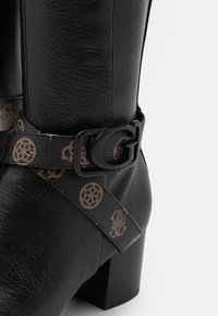 Bottine en cuir noir avec une finition texturée, un talon bloc, et une sangle à motif ornée d'une boucle noire affichant la lettre "G".