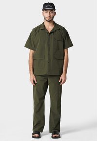 Camisa de manga corta en verde oliva con cierre de botones y dos bolsillos frontales, combinada con pantalones a juego y sandalias negras.