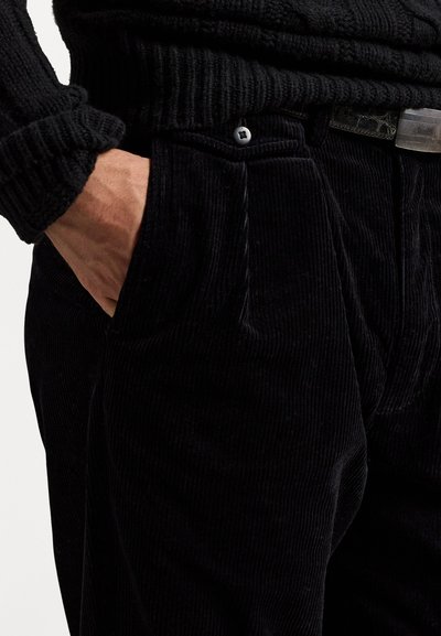 Pantalones de pana negros con costillas verticales, que cuentan con bolsillos frontales y una tela texturada. Una mano reposa de manera casual en un bolsillo.