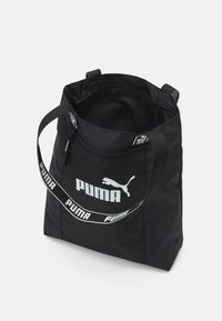 Sac fourre-tout noir Puma avec un grand logo blanc Puma, ouverture zippée et bandoulière de marque sur fond blanc.