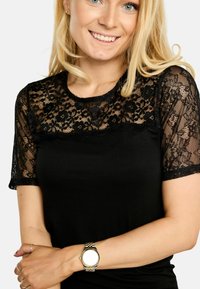 Top negru cu mâneci scurte din dantelă transparentă cu modele florale. Material fin pentru corp, cu decolteu rotund, purtat cu o curea de ceas metalică.