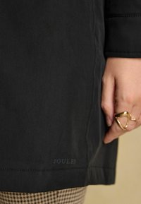 Schwarzer Stoffmantel mit glatter Textur, der am Saum ein dezentes "Joules"-Logo trägt. Mit einem goldenen Ring über karierter Hose handverziert.