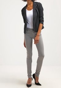 Harmaat slim-fit-housut yhdistettynä valkoiseen toppiin ja mustaan bleiseriin. Asuun kuuluu mustat stilettikorkokengät, ja sillä on sileä pinta.