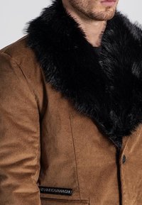 Chaqueta exterior marrón con un cuello de piel sintética negra, presenta una tela texturizada, cierre de botones y una etiqueta en el lado.