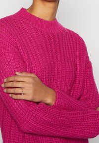 Neonpinkistä neulottu villapaita, jossa on ribbineulospinta ja pyöreä pääntie. Mallin kädessä näkyy hopeinen sormus.