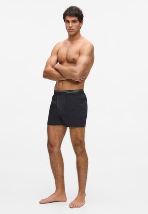 Homme musclé debout pieds nus, bras croisés, portant un boxer noir Karl Lagerfeld sur fond blanc uni.