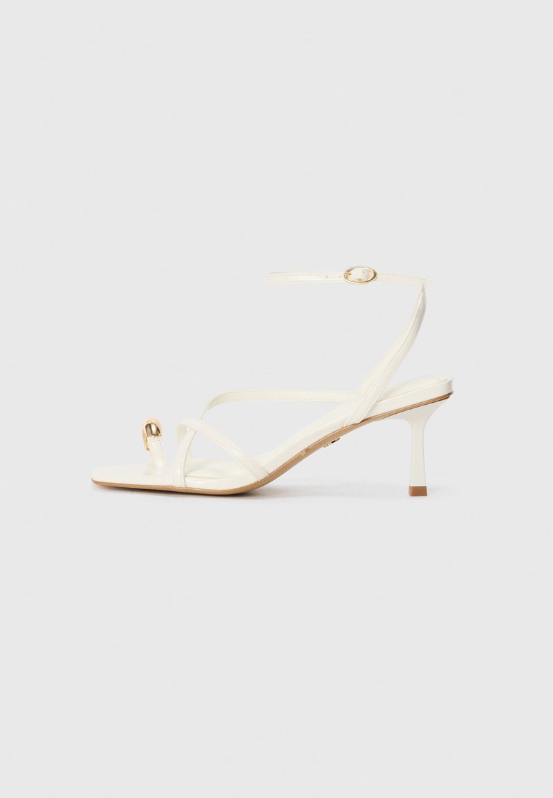 Hvide strophæle sandaler med spidse tæer, delikat ankelrem, og detaljer i guldfarvet metal. Glat tekstur og minimalistisk design.