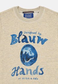 Sv vienkovej béžovej mikiny so modrým grafickým textom "Navrhnuté Blau Hands" a modrým kruhovým dizajnom. Mäkká bavlnená zmesová látka.