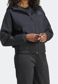 Svart zip-up hoodie i slät tyg med huva, framfickor och randiga detaljer på ärmarna. Matchas med svarta byxor.