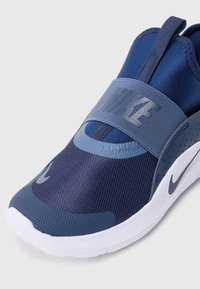 Chaussure de sport bleu marine avec une tige en matière textile, une sangle élastique et une semelle blanche. Présente une finition texturée et un logo de couleur contrastante.