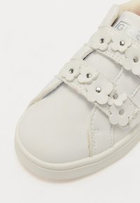 Zapatilla blanca con acentos florales, material de cuero suave, tres cierres de tira y herrajes plateados redondos. Punta redondeada y suela plana.