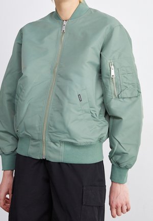Personne portant une veste bomber vert clair avec fermeture éclair frontale et poche zippée sur la manche, associée à un pantalon cargo noir.