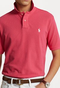 Röd pikétröja med slim fit och kort krage, med en vit broderad logotyp på vänster bröst och textilstruktur. Brun bälte och klocka synliga.