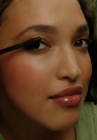 Svart mascara borste som appliceras på långa, lockiga ögonfransar. Slät, glödande hy med en mjuk rosa rouge och glänsande nude läppar. Grön bakgrund.