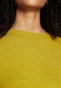 Gros plan sur le cou et les épaules d'une personne aux cheveux bouclés foncés portant un haut texturé jaune-vert vif et une petite boucle d'oreille.