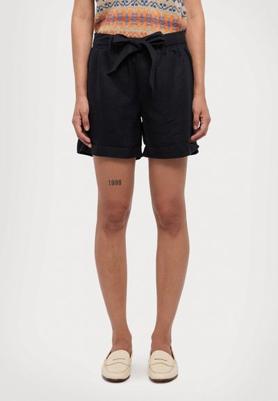 Shorts din in cuiva marin, cu un cordon la talie, manșete răsucite și buzunare laterale; modelul prezintă o tatuaj vizibil pe coapsa dreaptă.