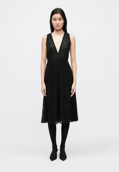Black pleated dress with a deep V-neck and sleeveless design, featuring shimmering accents and a fitted waist, paired with black tights.