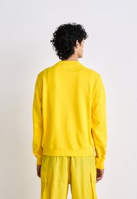 Felpa gialla con collo rotondo e maniche lunghe, realizzata in materiale morbido. Presenta un orlo e polsini a costine, indossata con pantaloni abbinati.