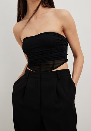 Kvinde iført en sort stropløs rynket cropped top med gennemsigtigt mesh panel kombineret med højtaljede sorte bukser, hænderne i lommerne.