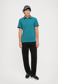 Teal polo skjorte med et mønstrete svart krage og ermer, kombinert med svarte bukser og en grå caps. Sneakers fullfører antrekket.