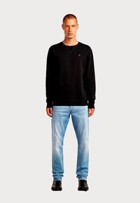 Felpa nera con scollo rotondo, polsini a coste e un piccolo logo sul petto, abbinata a jeans azzurri e stivali neri.