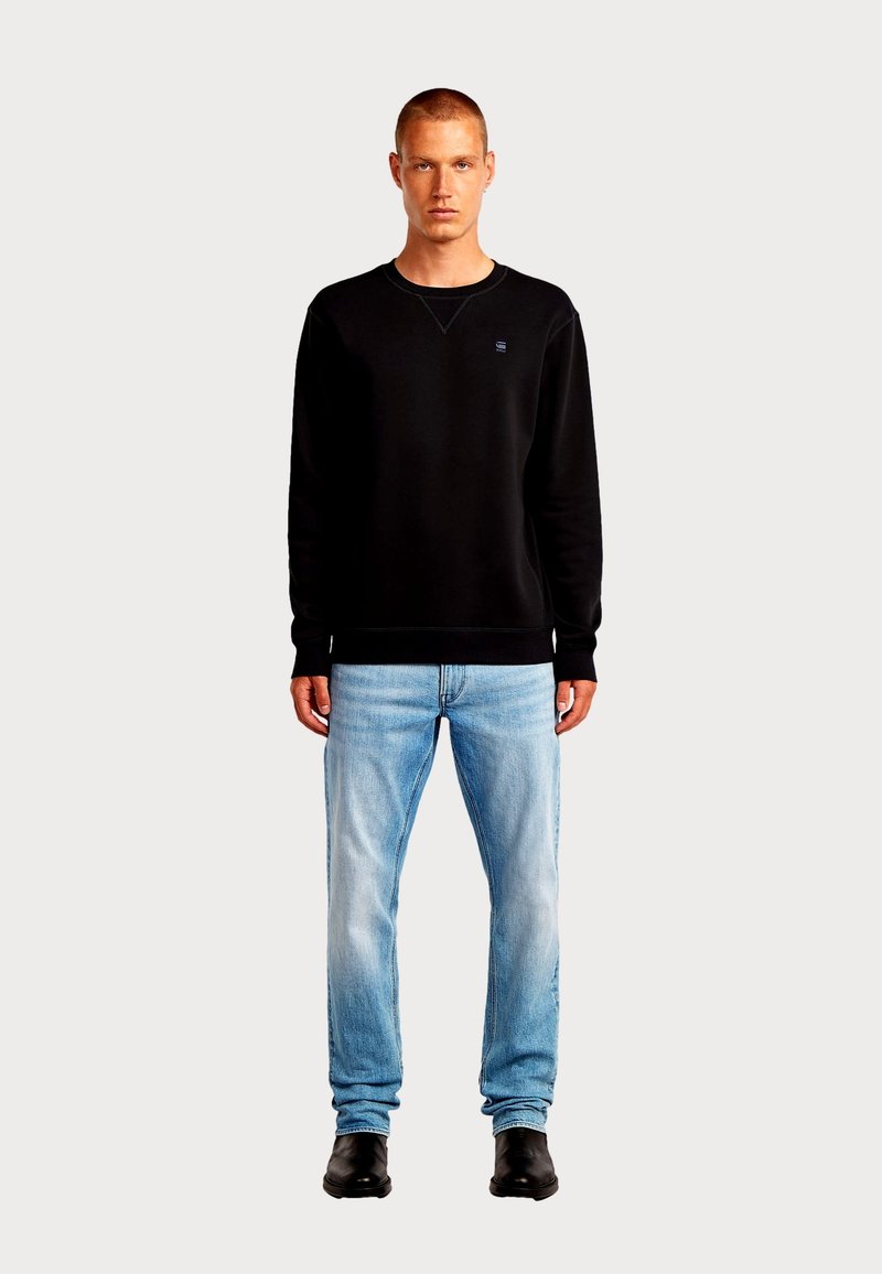 Sweat-shirt noir avec col rond, poignets en maille côtelée et un petit logo sur la poitrine, accompagné d'un jean bleu clair et de bottes noires.