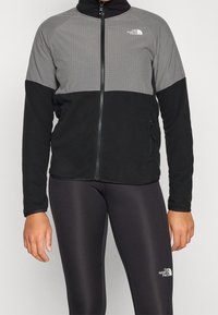 Giacca in pile con zip frontale, parte superiore grigia a trama a griglia e parte inferiore nera. Include una tasca laterale e un accento con logo bianco.