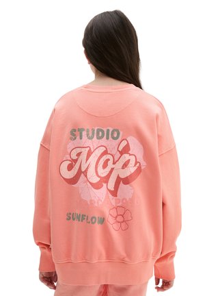 Person mit langen dunklen Haaren, die einen übergroßen pastellkorallenfarbenen Pullover mit dem Schriftzug „STUDIO Mop SUNFLOW“ und einem Blumendesign auf dem Rücken trägt.