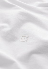 T-shirt bianco in cotone con una texture liscia, caratterizzato da un logo sottile al centro. Tessuto morbido e leggero che si adagia dolcemente.