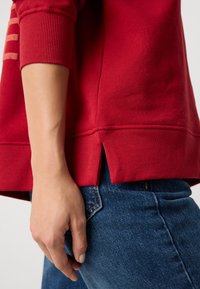 Roter Sweatshirt mit gerippten Bündchen und Saum, versehen mit seitlichen Schlitzen. Kombiniert mit dunkelblauen Jeans, die eine Nahaufnahme der Textur des Stoffes zeigen.
