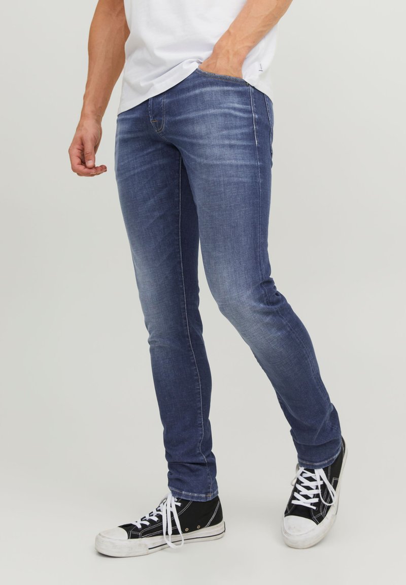 Jean en denim bleu avec une coupe slim, léger délavage, design à cinq poches, et coutures beige, associés à des chaussures de sport noires montantes.