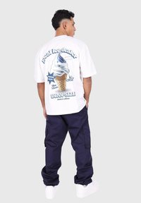 T-shirt en coton blanc avec un graphique de cornet de glace et du texte bleu au dos, associé à un pantalon cargo bleu marine et des baskets blanches. Style décontracté urbain.