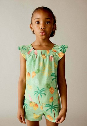 Jeune fille portant une tenue verte avec des motifs de fruits tropicaux et de palmiers, debout devant un fond uni de couleur claire.